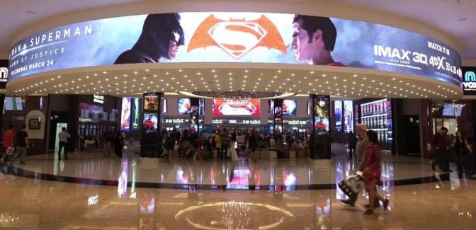A bustling shopping mall with people strolling around, featuring a large screen displaying movies in a Saudi Arabian movie salon.