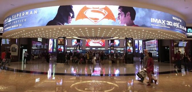 A bustling shopping mall with people strolling around, featuring a large screen displaying movies in a Saudi Arabian movie salon.
