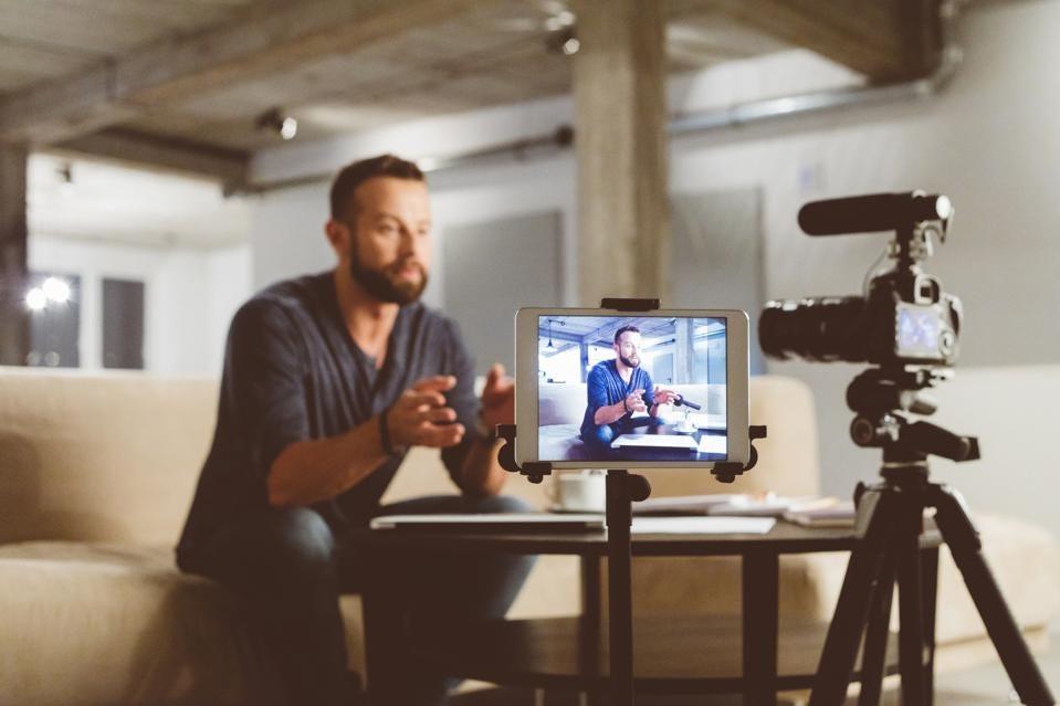 A man in casual clothing records a video blog, speaking to a camera mounted on a tripod with a screen displaying his image in a softly lit indoor setting.