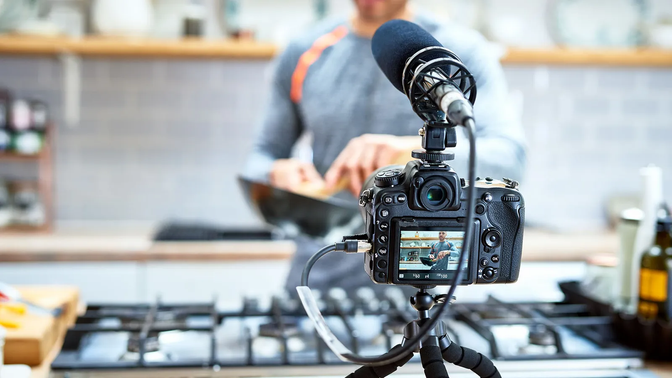 A digital camera on a tripod records a cooking demonstration. the camera screen displays a person in an apron at a stove, stirring a pan. focus is on the camera with a blurry kitchen background.