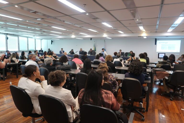 Sala ampla de audiência ou seminário, com diversas pessoas sentadas em mesas voltadas para uma mesa principal ao fundo. No painel frontal, participantes acompanham a apresentação enquanto um conteúdo é projetado em uma tela à direita. O ambiente é institucional, com iluminação clara e público formado por profissionais reunidos para debate ou apresentação.
