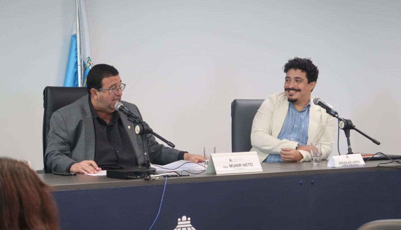 Dois homens estão sentados à mesa durante uma audiência ou reunião institucional, ambos diante de microfones. À esquerda, um homem de terno fala enquanto consulta documentos; à direita, outro homem, de blazer claro e camisa azul, sorri enquanto o observa. Sobre a mesa, placas identificam “Dep. Munir Neto” e “Douglas Lopes”. Ao fundo, há uma bandeira e, na parte frontal da mesa, o símbolo da Assembleia Legislativa do Estado do Rio de Janeiro.