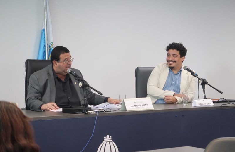 Dois homens estão sentados à mesa durante uma audiência ou reunião institucional, ambos diante de microfones. À esquerda, um homem de terno fala enquanto consulta documentos; à direita, outro homem, de blazer claro e camisa azul, sorri enquanto o observa. Sobre a mesa, placas identificam “Dep. Munir Neto” e “Douglas Lopes”. Ao fundo, há uma bandeira e, na parte frontal da mesa, o símbolo da Assembleia Legislativa do Estado do Rio de Janeiro.