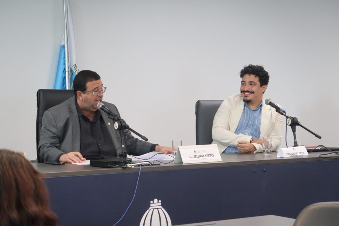 Dois homens estão sentados à mesa durante uma audiência ou reunião institucional, ambos diante de microfones. À esquerda, um homem de terno fala enquanto consulta documentos; à direita, outro homem, de blazer claro e camisa azul, sorri enquanto o observa. Sobre a mesa, placas identificam “Dep. Munir Neto” e “Douglas Lopes”. Ao fundo, há uma bandeira e, na parte frontal da mesa, o símbolo da Assembleia Legislativa do Estado do Rio de Janeiro.