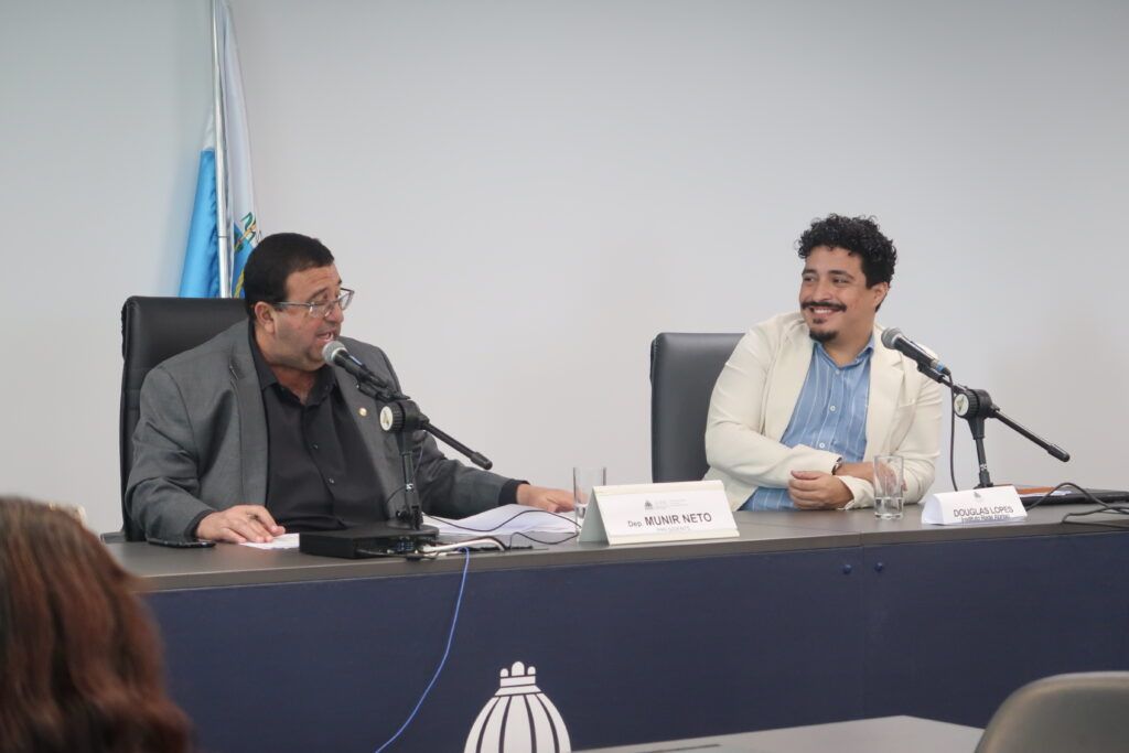 Dois homens estão sentados à mesa durante uma audiência ou reunião institucional, ambos diante de microfones. À esquerda, um homem de terno fala enquanto consulta documentos; à direita, outro homem, de blazer claro e camisa azul, sorri enquanto o observa. Sobre a mesa, placas identificam “Dep. Munir Neto” e “Douglas Lopes”. Ao fundo, há uma bandeira e, na parte frontal da mesa, o símbolo da Assembleia Legislativa do Estado do Rio de Janeiro.