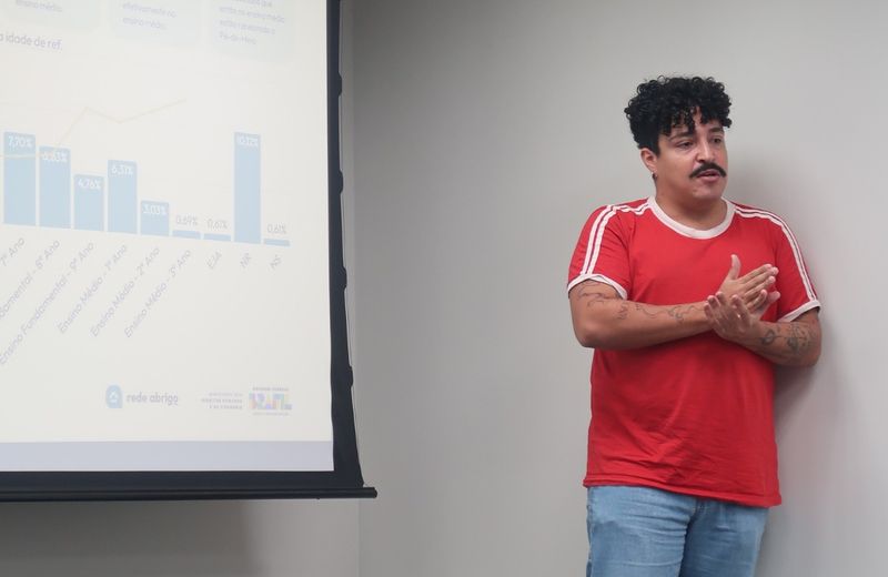 Um homem de camiseta vermelha apresenta dados em uma sala, posicionado ao lado de uma tela de projeção com gráficos e percentuais sobre acolhimento. Ele gesticula enquanto fala, em um ambiente de reunião ou apresentação institucional.