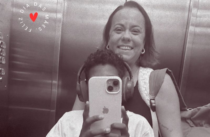 Foto em preto e branco feita diante do espelho de um elevador. Uma criança, usando fones de ouvido, segura um celular à frente do rosto para registrar a imagem, enquanto uma mulher sorridente aparece logo atrás. No canto superior esquerdo, há um coração vermelho cercado pela frase “Feliz Dia das Mães”.