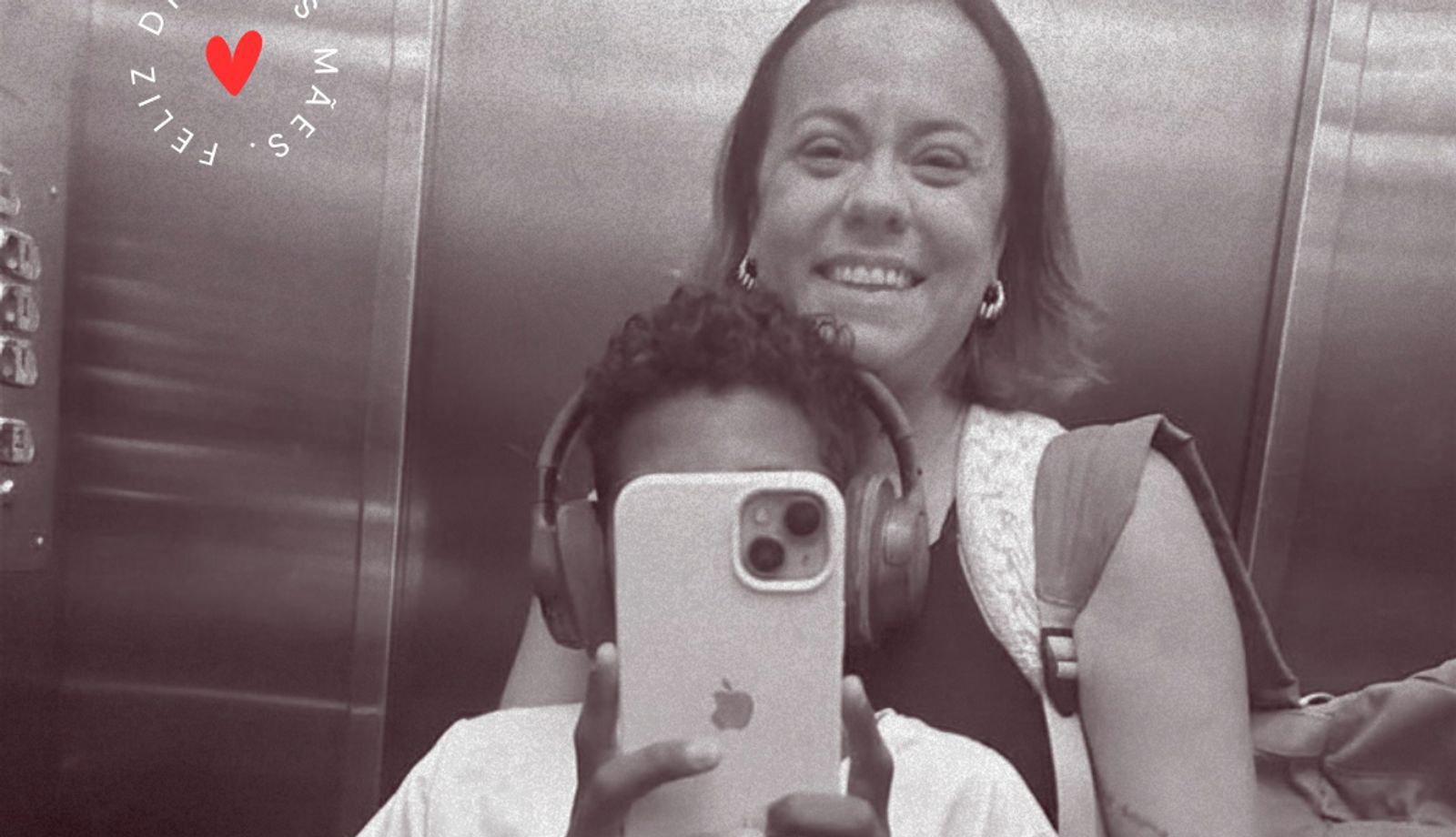 Foto em preto e branco feita diante do espelho de um elevador. Uma criança, usando fones de ouvido, segura um celular à frente do rosto para registrar a imagem, enquanto uma mulher sorridente aparece logo atrás. No canto superior esquerdo, há um coração vermelho cercado pela frase “Feliz Dia das Mães”.
