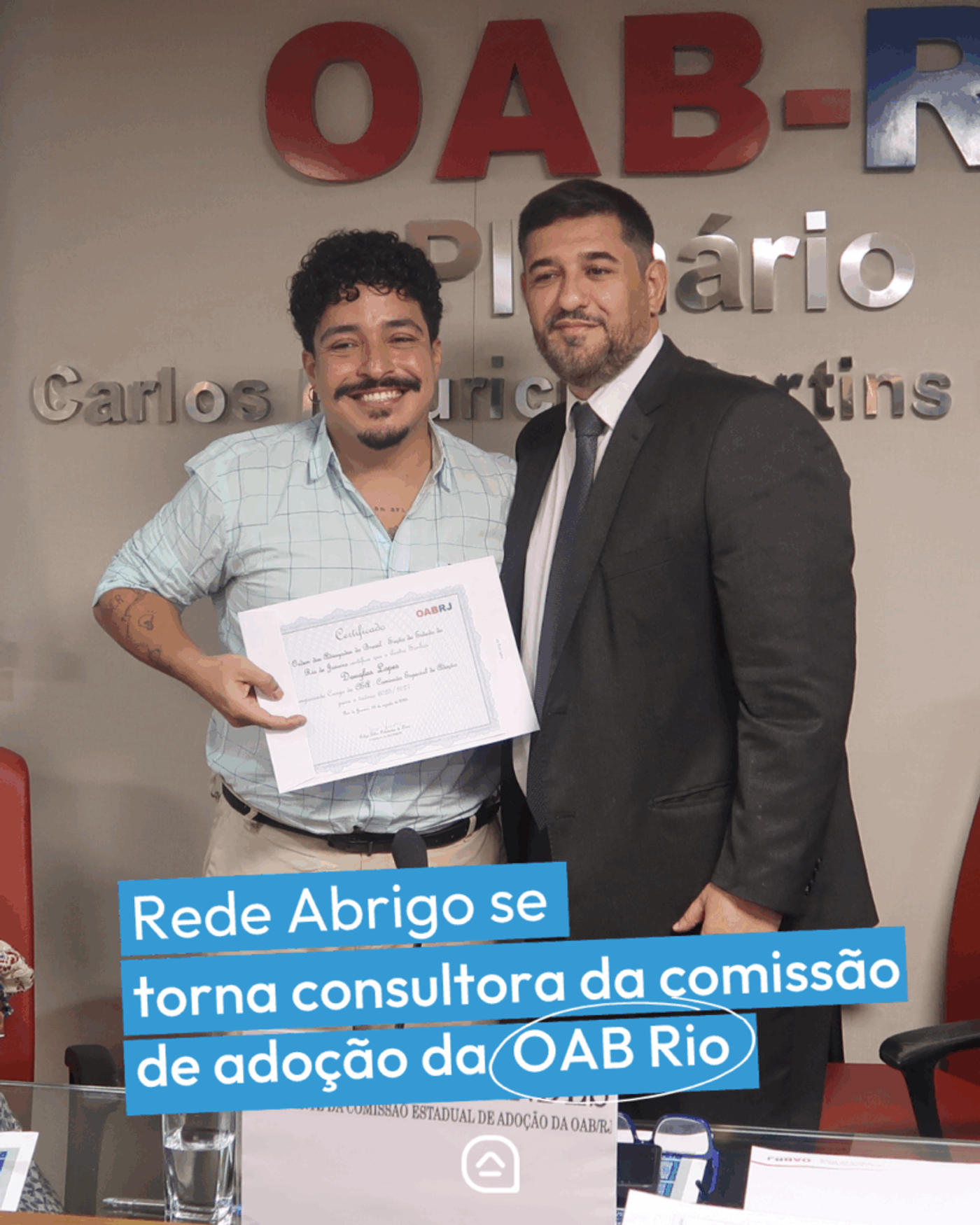 Dois homens posam lado a lado em frente ao painel da OAB-RJ. Um deles segura um certificado enquanto o outro, de terno e gravata, está ao seu lado. Na parte inferior da imagem, aparece a frase: “Rede Abrigo se torna consultora da comissão de adoção da OAB Rio”.