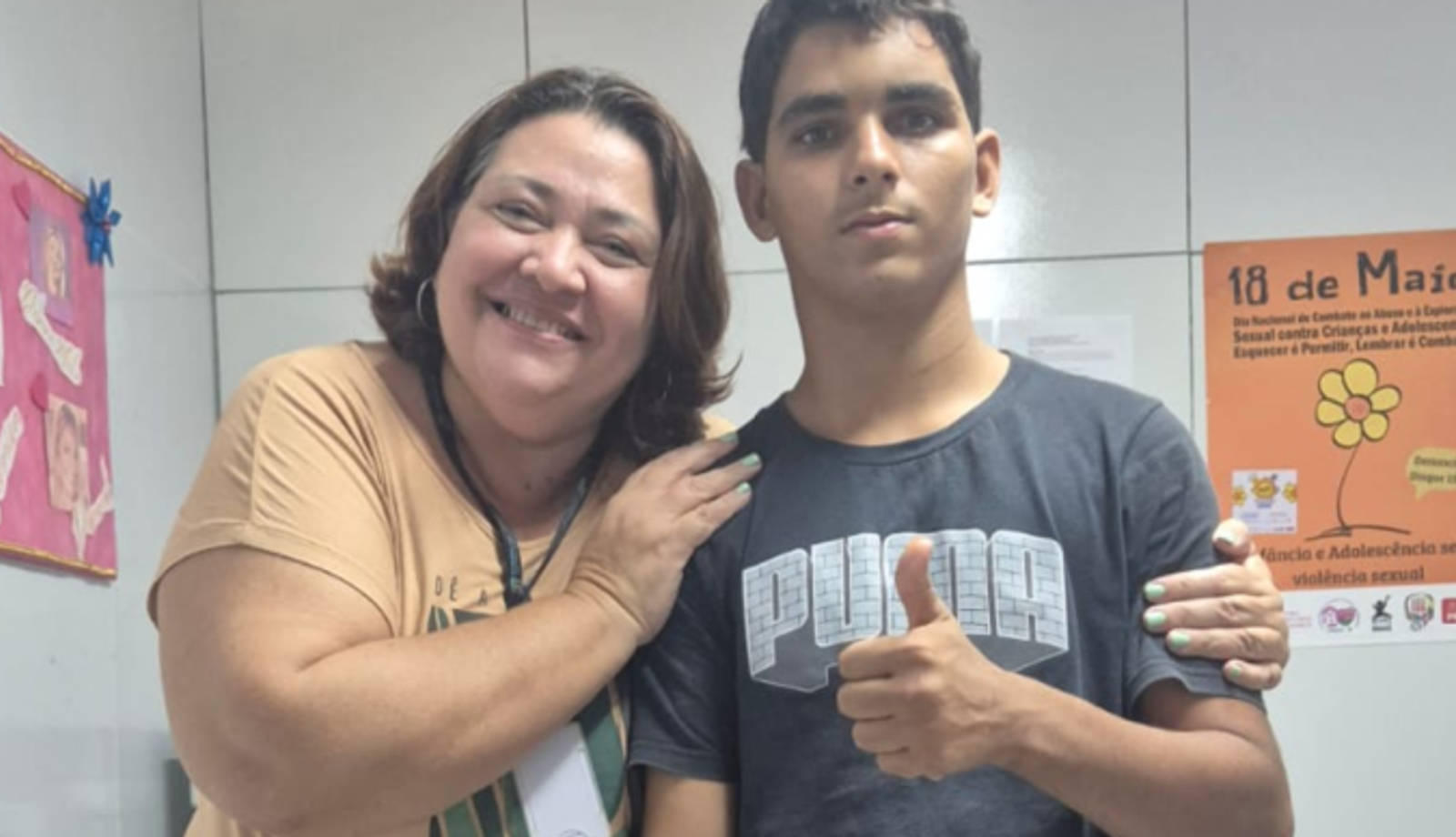 Uma mulher sorri ao lado de um adolescente, com o braço apoiado sobre o ombro dele. O jovem veste camiseta escura e faz sinal de positivo com a mão. Ao fundo, há cartazes informativos sobre direitos de crianças e adolescentes e prevenção à violência sexual.