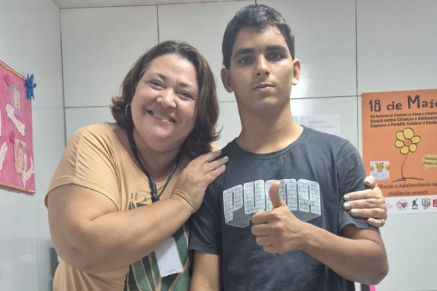 Uma mulher sorri ao lado de um adolescente, com o braço apoiado sobre o ombro dele. O jovem veste camiseta escura e faz sinal de positivo com a mão. Ao fundo, há cartazes informativos sobre direitos de crianças e adolescentes e prevenção à violência sexual.