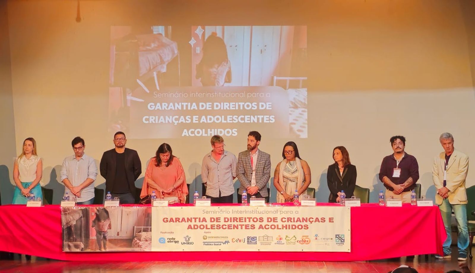 Grupo de participantes está em pé atrás da mesa principal durante o Seminário Interinstitucional para a Garantia de Direitos de Crianças e Adolescentes Acolhidos. Ao fundo, uma projeção exibe o nome do evento, e a mesa está identificada com faixa institucional e logotipos das organizações participantes.