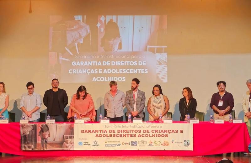 Grupo de participantes está em pé atrás da mesa principal durante o Seminário Interinstitucional para a Garantia de Direitos de Crianças e Adolescentes Acolhidos. Ao fundo, uma projeção exibe o nome do evento, e a mesa está identificada com faixa institucional e logotipos das organizações participantes.