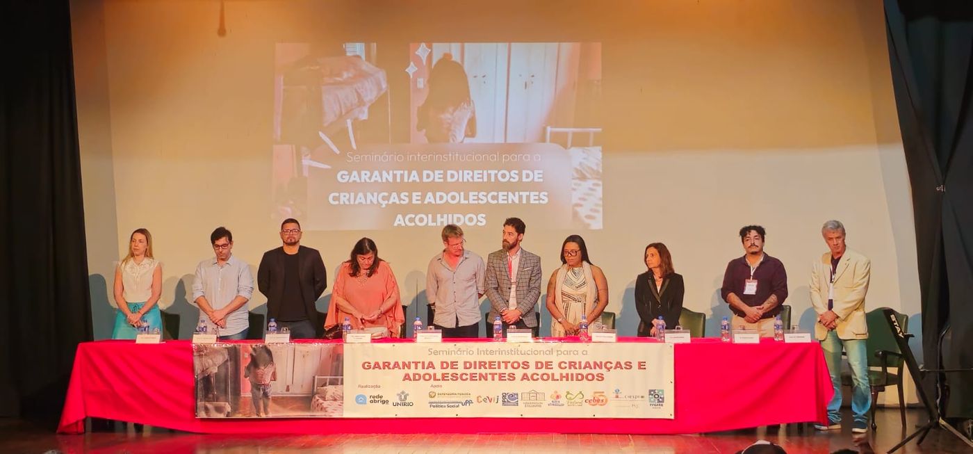 Grupo de participantes está em pé atrás da mesa principal durante o Seminário Interinstitucional para a Garantia de Direitos de Crianças e Adolescentes Acolhidos. Ao fundo, uma projeção exibe o nome do evento, e a mesa está identificada com faixa institucional e logotipos das organizações participantes.