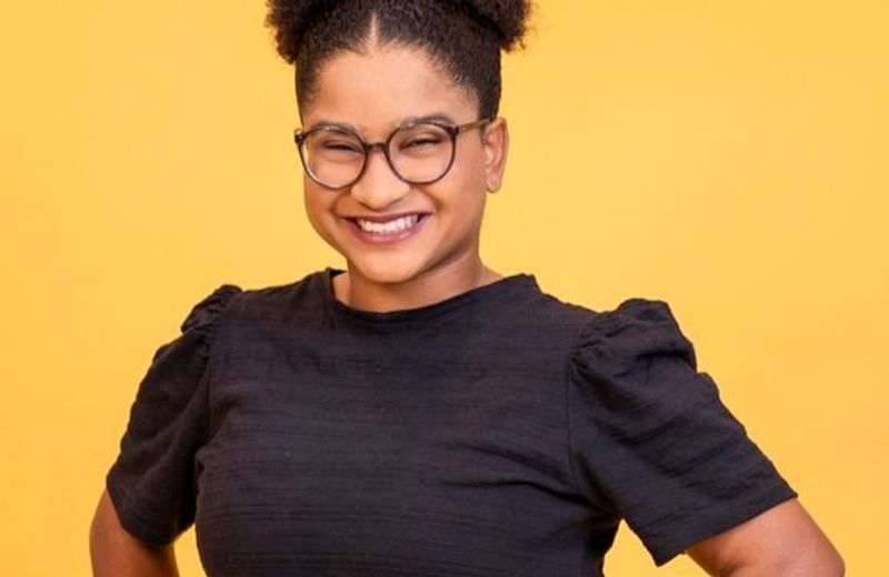 Retrato de uma mulher sorrindo diante de um fundo amarelo. Ela usa óculos de armação escura, cabelo preso em um coque alto e volumoso e veste uma blusa preta de mangas curtas. A pose, com as mãos na cintura, transmite confiança e simpatia.