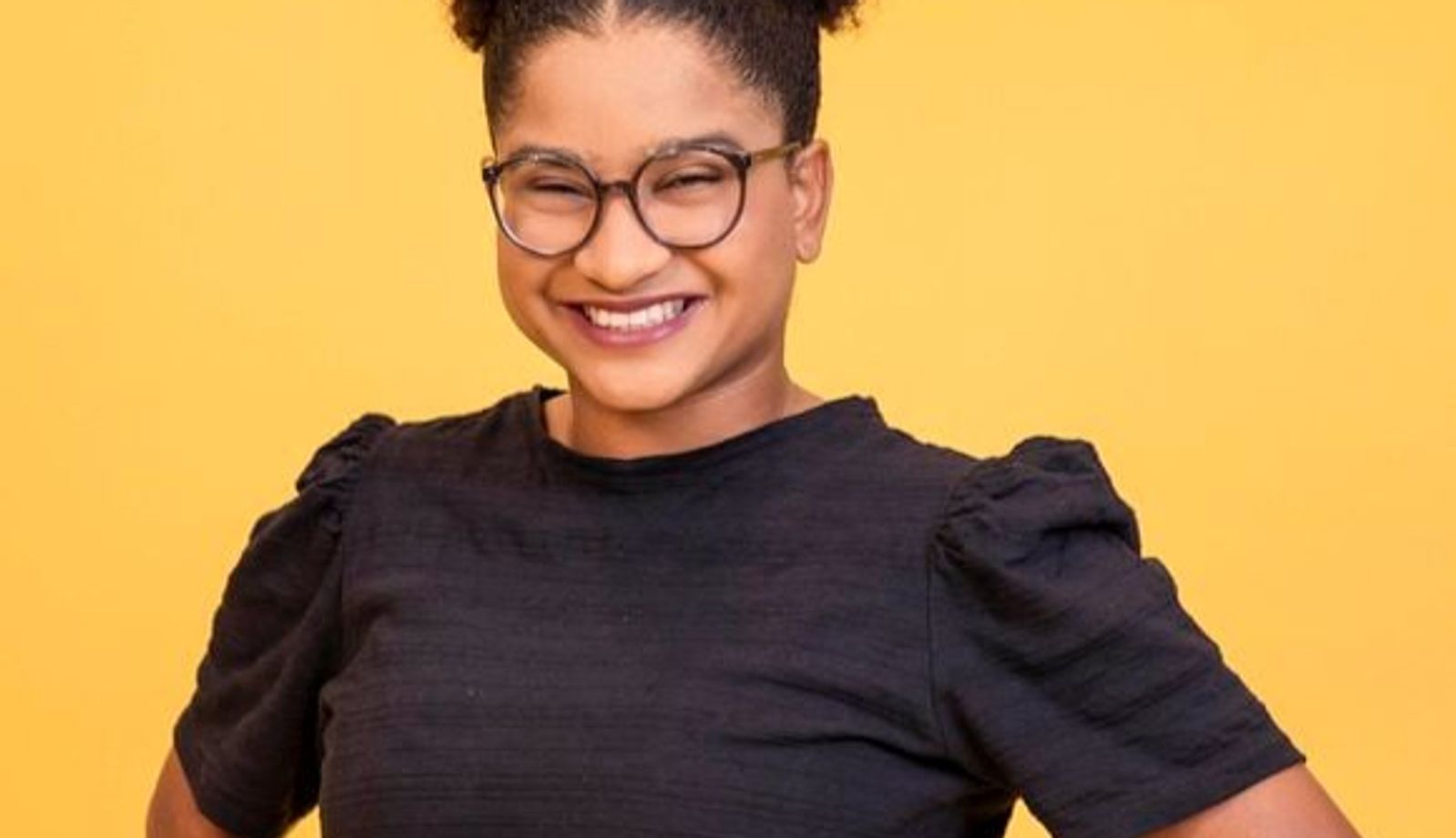 Retrato de uma mulher sorrindo diante de um fundo amarelo. Ela usa óculos de armação escura, cabelo preso em um coque alto e volumoso e veste uma blusa preta de mangas curtas. A pose, com as mãos na cintura, transmite confiança e simpatia.