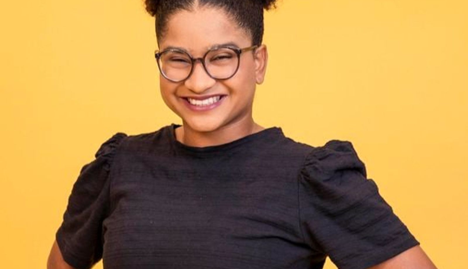 Retrato de uma mulher sorrindo diante de um fundo amarelo. Ela usa óculos de armação escura, cabelo preso em um coque alto e volumoso e veste uma blusa preta de mangas curtas. A pose, com as mãos na cintura, transmite confiança e simpatia.