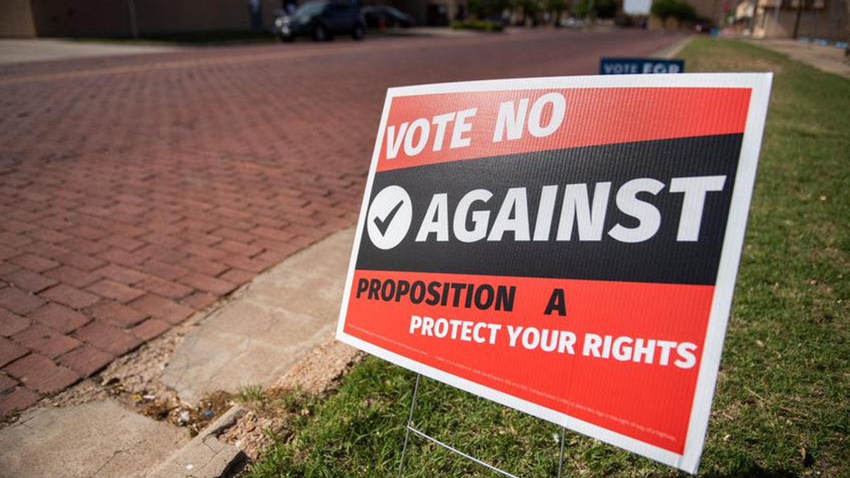 A red and black sign posted near a sidewalk that reads: "Vote NO Against Proposition A Protect your Rights"