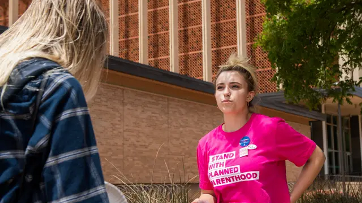 Blond woman wearing a pink Planned Parenthood shirt that reads "Stand with Planned Parenthood" is talking to another person with long blond hair at a table. 