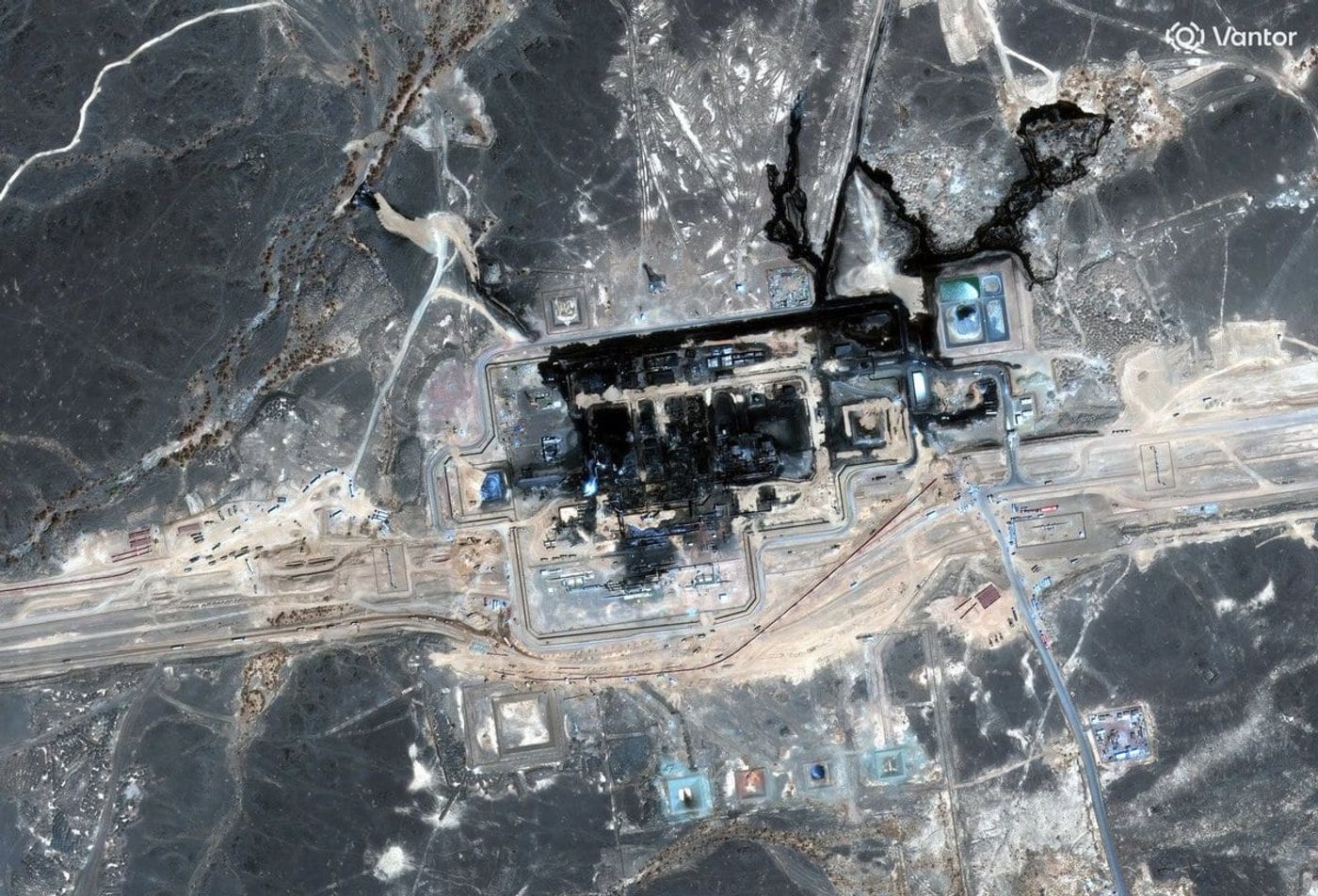 Satellite view of a desert pumping station after a strike: the whole walled process area scorched black, and a large slick of spilled crude spreading across open ground beyond the northeast corner of the perimeter.