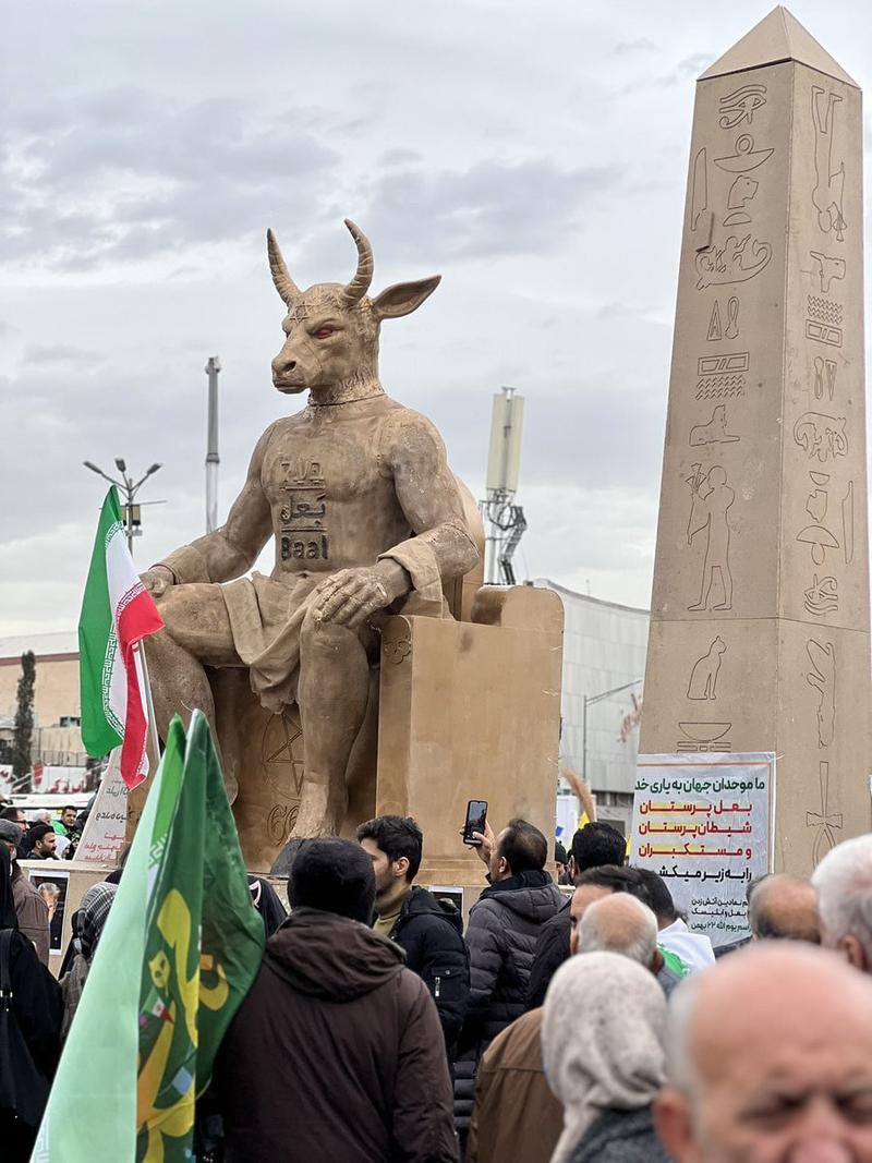 A street level photograph taken during a public demonstration. In the foreground, a large, tan-colored statue of a bull-headed humanoid figure sits on a throne; the word "Baal" is written on its chest in English and Arabic. To the right of the figure stands a tall obelisk covered in faux hieroglyphs. In the background, a crowd of people holds green, white, and red Iranian flags.