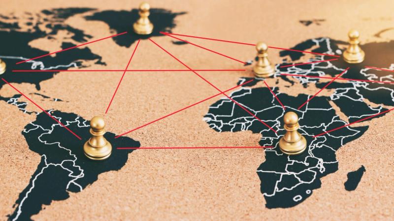 A stylized black world map pinned to a corkboard background. Gold pawn-shaped markers are placed on North America, South America, Europe, Africa, and Asia. Red threads connect these markers to one another in a complex, distributed web, visually representing a decentralized global network.