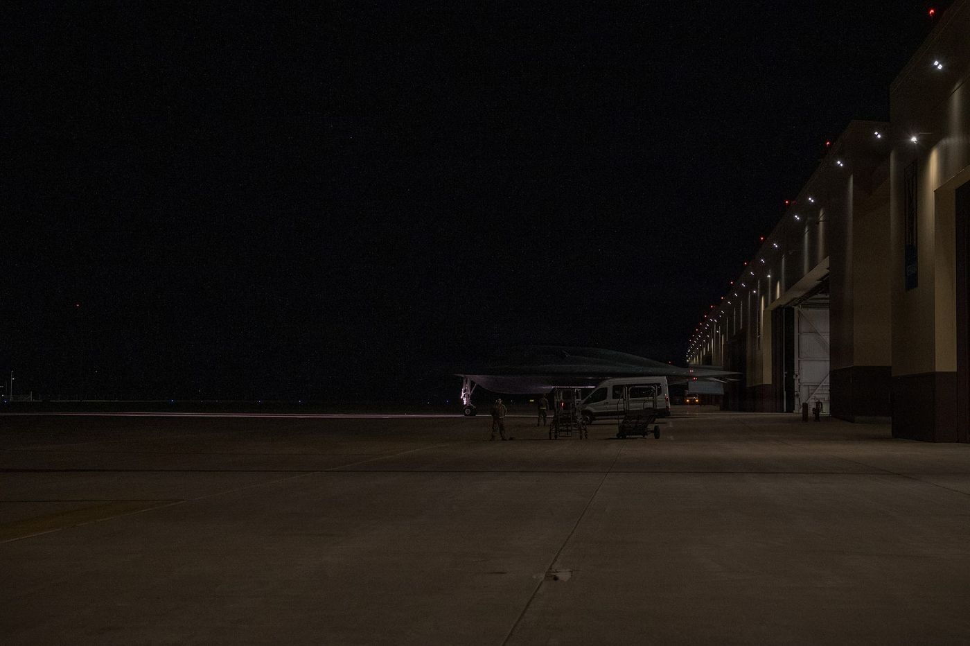 A B-2 Spirit on a darkened apron at night, lit hangars behind it