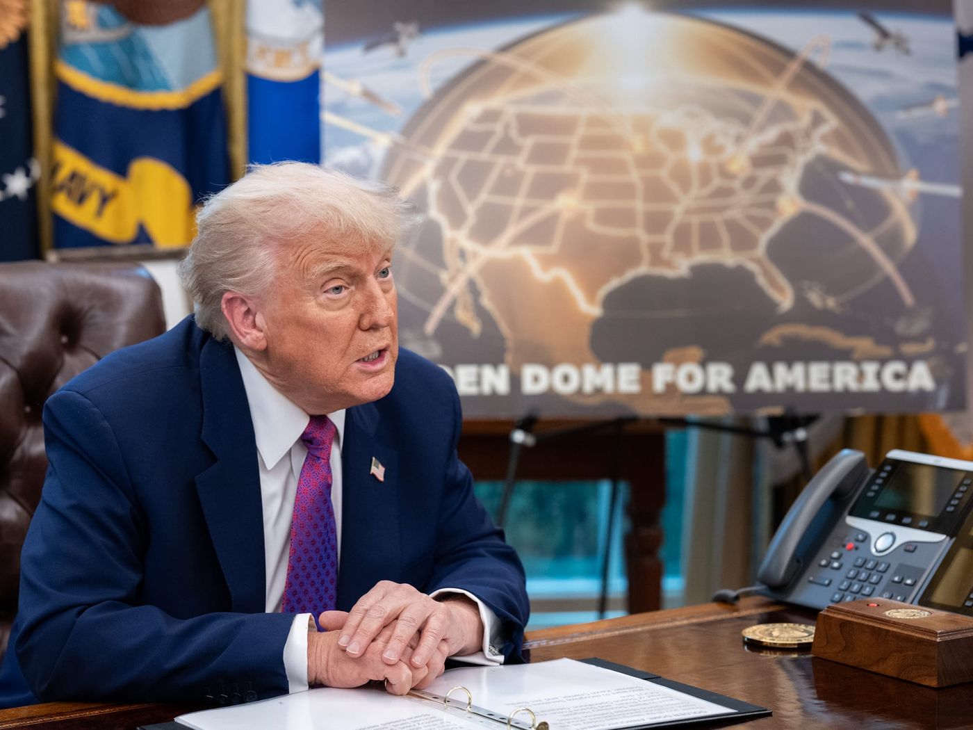 Donald Trump seated at the Resolute Desk with a Golden Dome for America graphic on the screen behind him