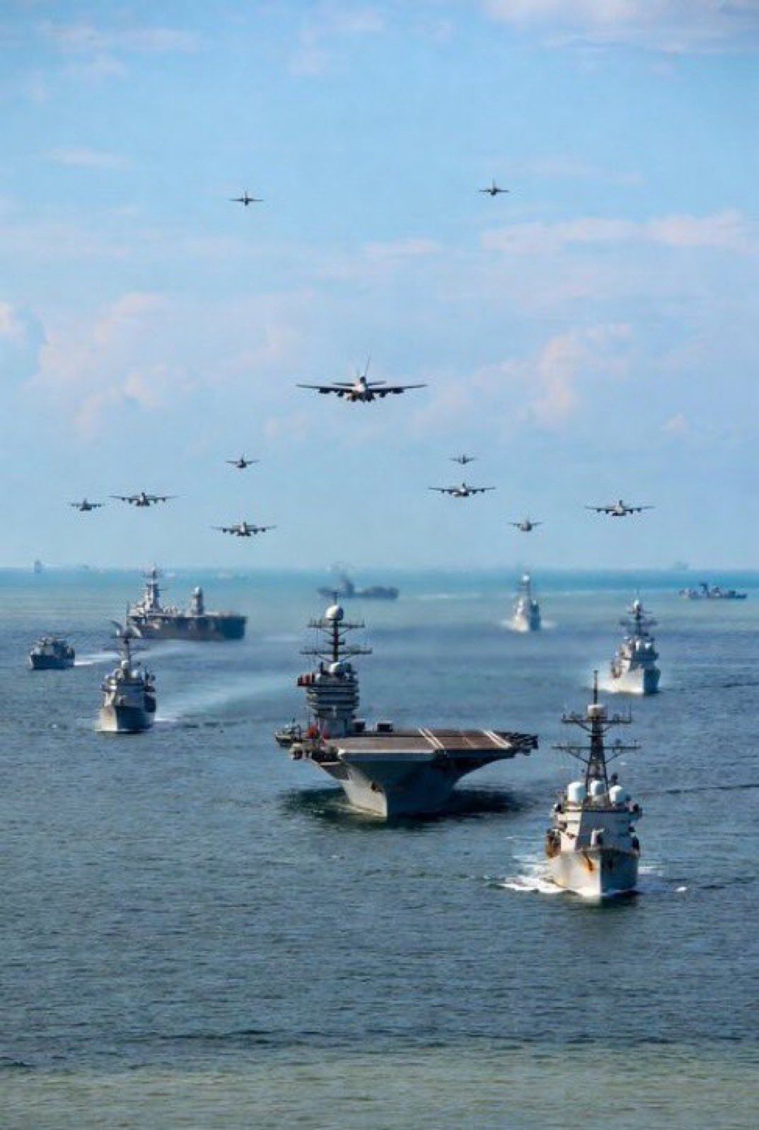 A large naval fleet, including an aircraft carrier, sails on the ocean as many military aircraft fly in formation overhead.