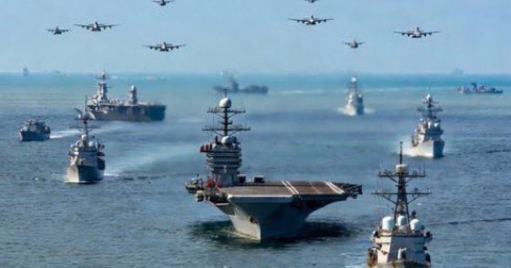 A large naval fleet, including an aircraft carrier, sails on the ocean as many military aircraft fly in formation overhead.