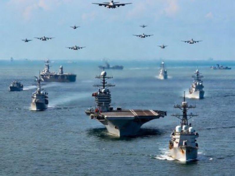 A large naval fleet, including an aircraft carrier, sails on the ocean as many military aircraft fly in formation overhead.