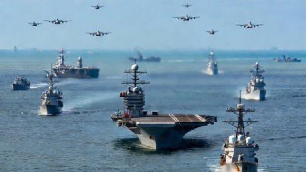 A large naval fleet, including an aircraft carrier, sails on the ocean as many military aircraft fly in formation overhead.