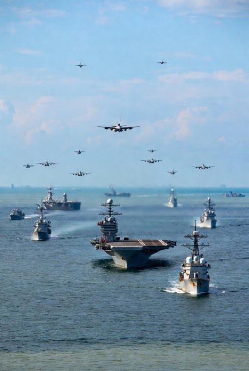 An aerial view of a U.S. Navy carrier strike group moving in formation on the blue ocean, with a squadron of fighter jets and bombers flying in formation overhead.