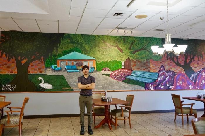 A wide angle shot of Manuel standing in front of a mural in a restaurant 
