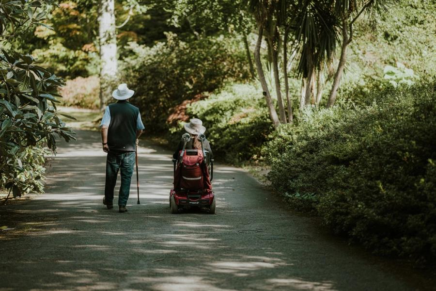 Two people moving along a shaded garden path, one walking with a cane and one using a mobility scooter with crutches stowed at the back.