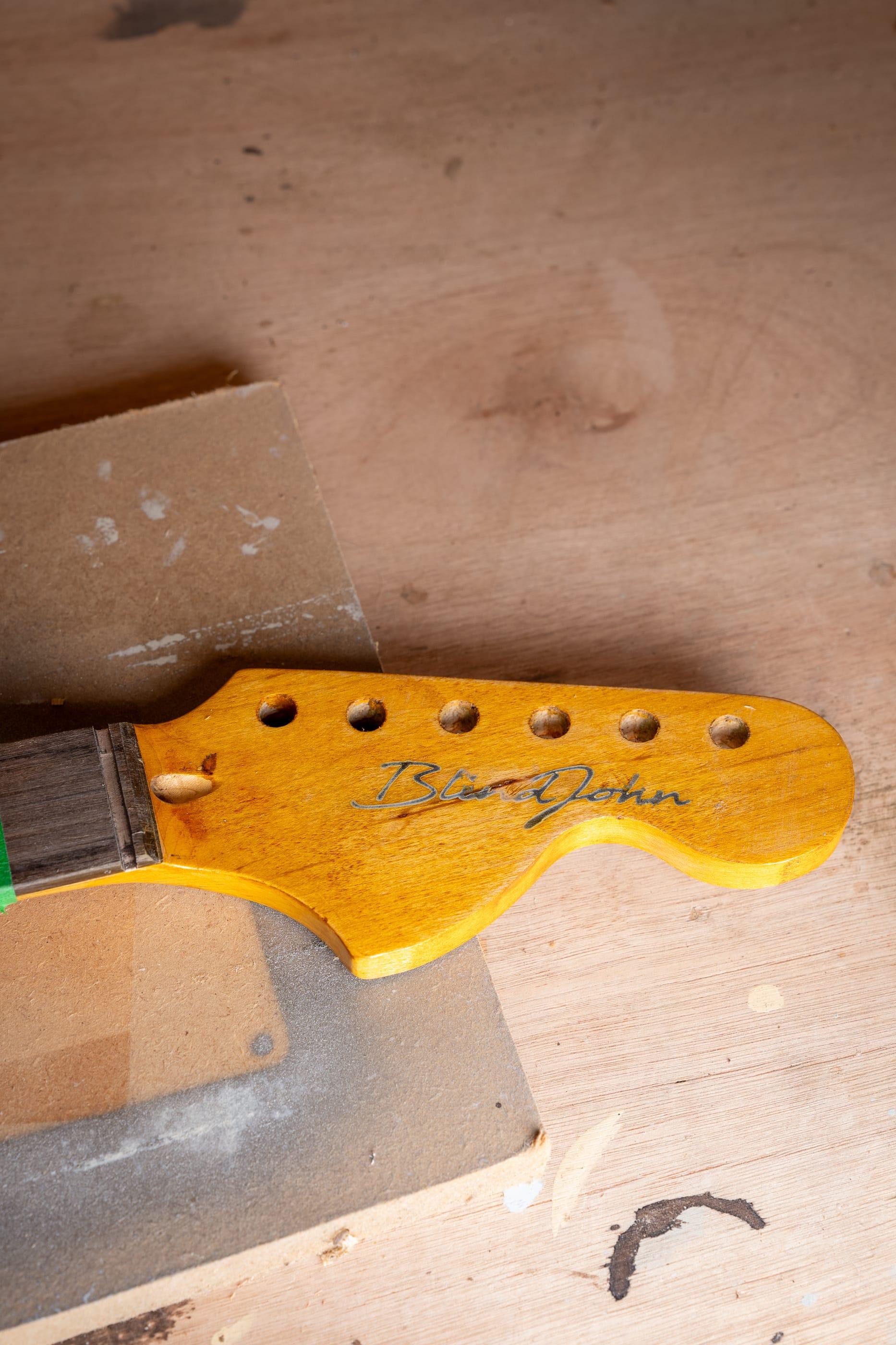 An unfinished guitar neck on the bench, the Blind John wordmark on its headstock