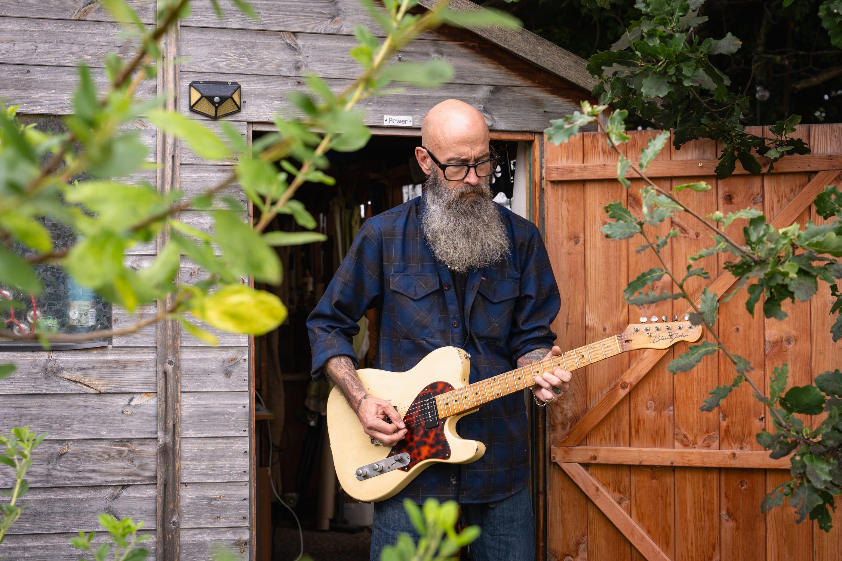 John playing a guitar in the open doorway of the shed, the oak in front