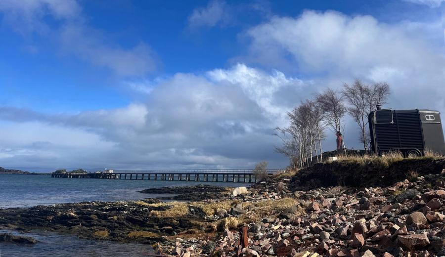 The wild sauna on the rocky shore beside the pier, birch trees behind and blue sky over the water
