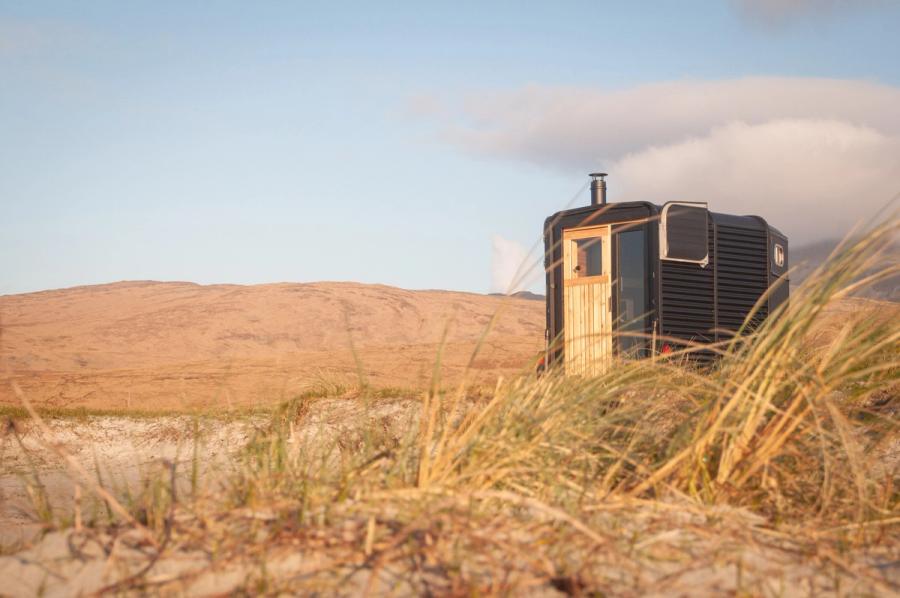 The converted horsebox sauna in marram grass at golden hour, smoke rising from its flue