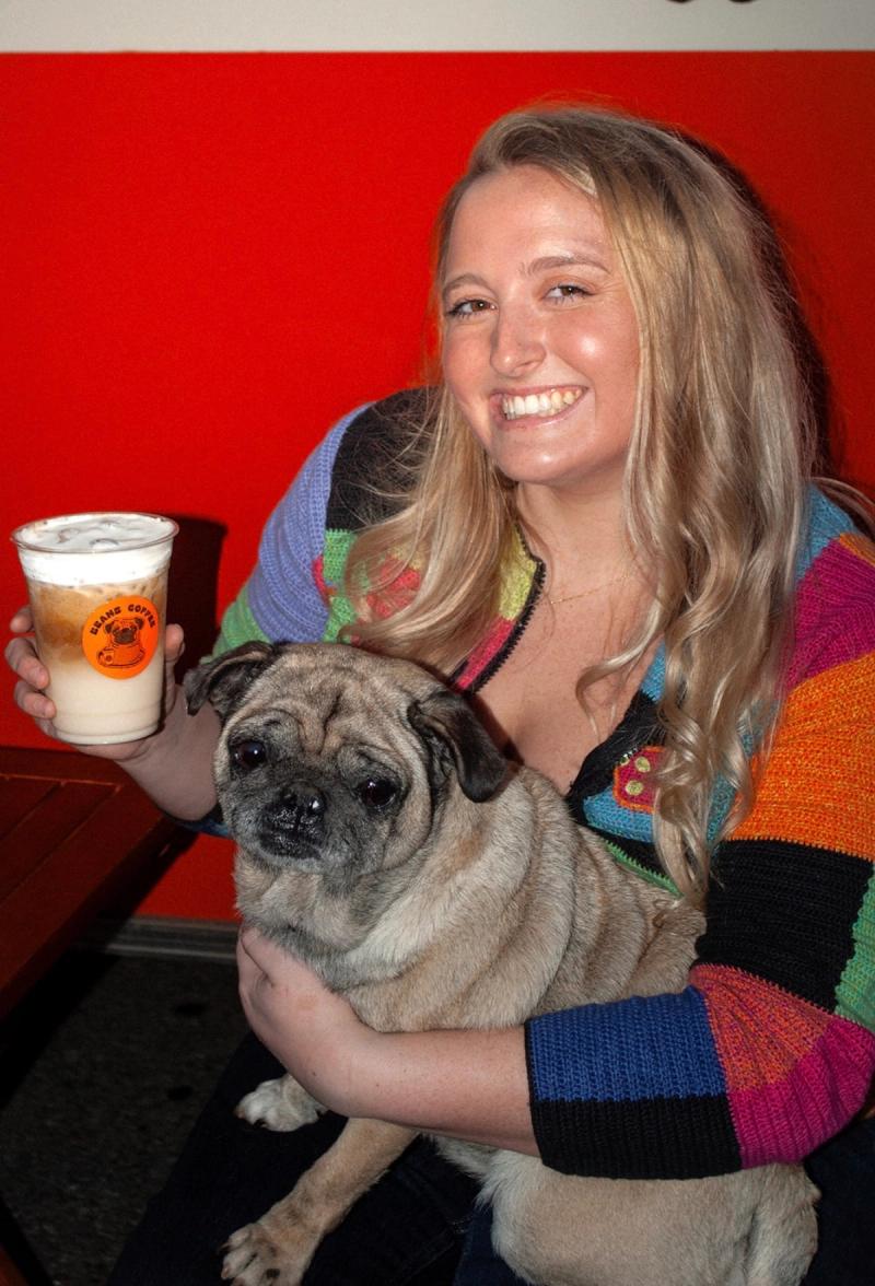 Cassidy on a bench with the pug on her lap, holding up an iced latte