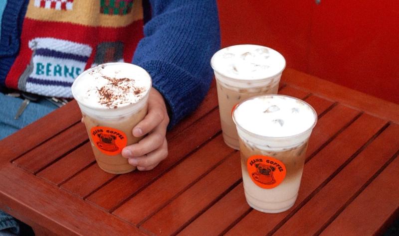 Three iced lattes on the wooden table, one of them held by a hand in a knitted Beans sweater