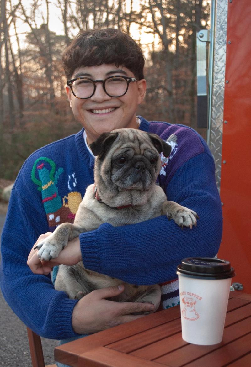 Brian in a blue knit sweater holding the pug at an outdoor table, a hot cup with the pug logo next to them