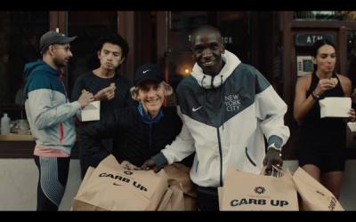 Eliud Kipchoge and Joan Benoit Samuelsson stand in front of a restaurant with takeout bags. 
