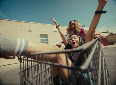 Two teens scurry through a parking lot in a shopping cart