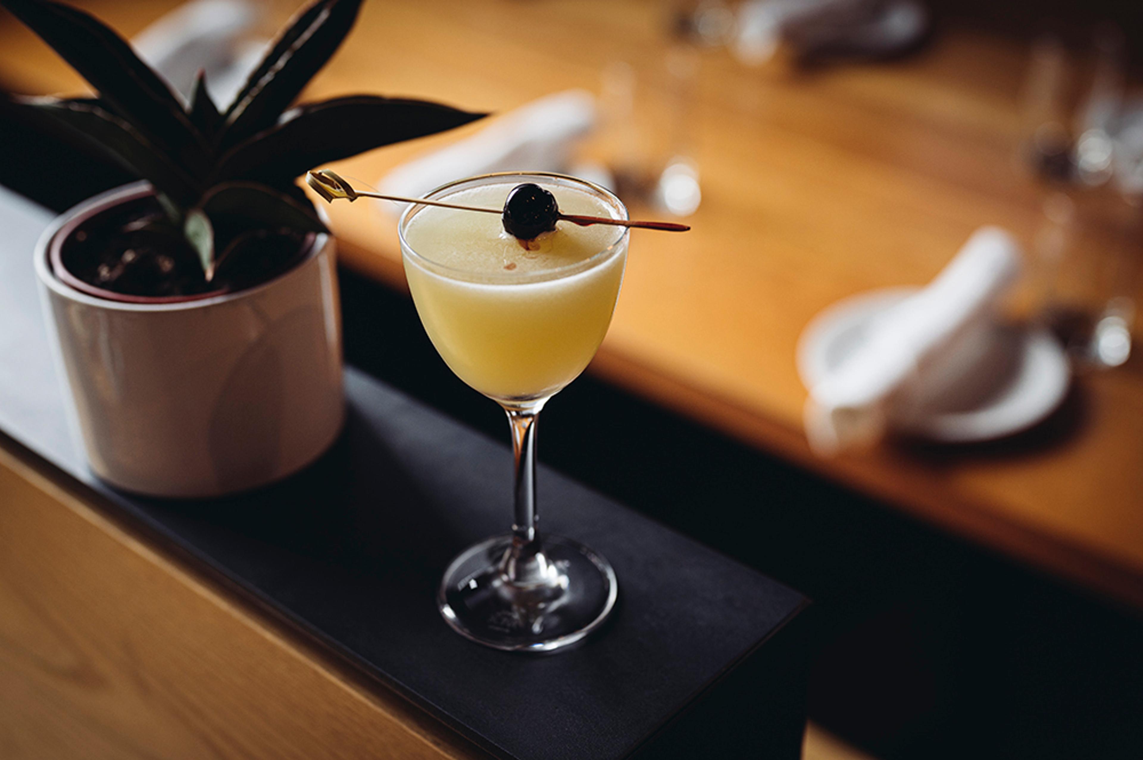 pale yellow cocktail in stem glass with brandied cherry garnish