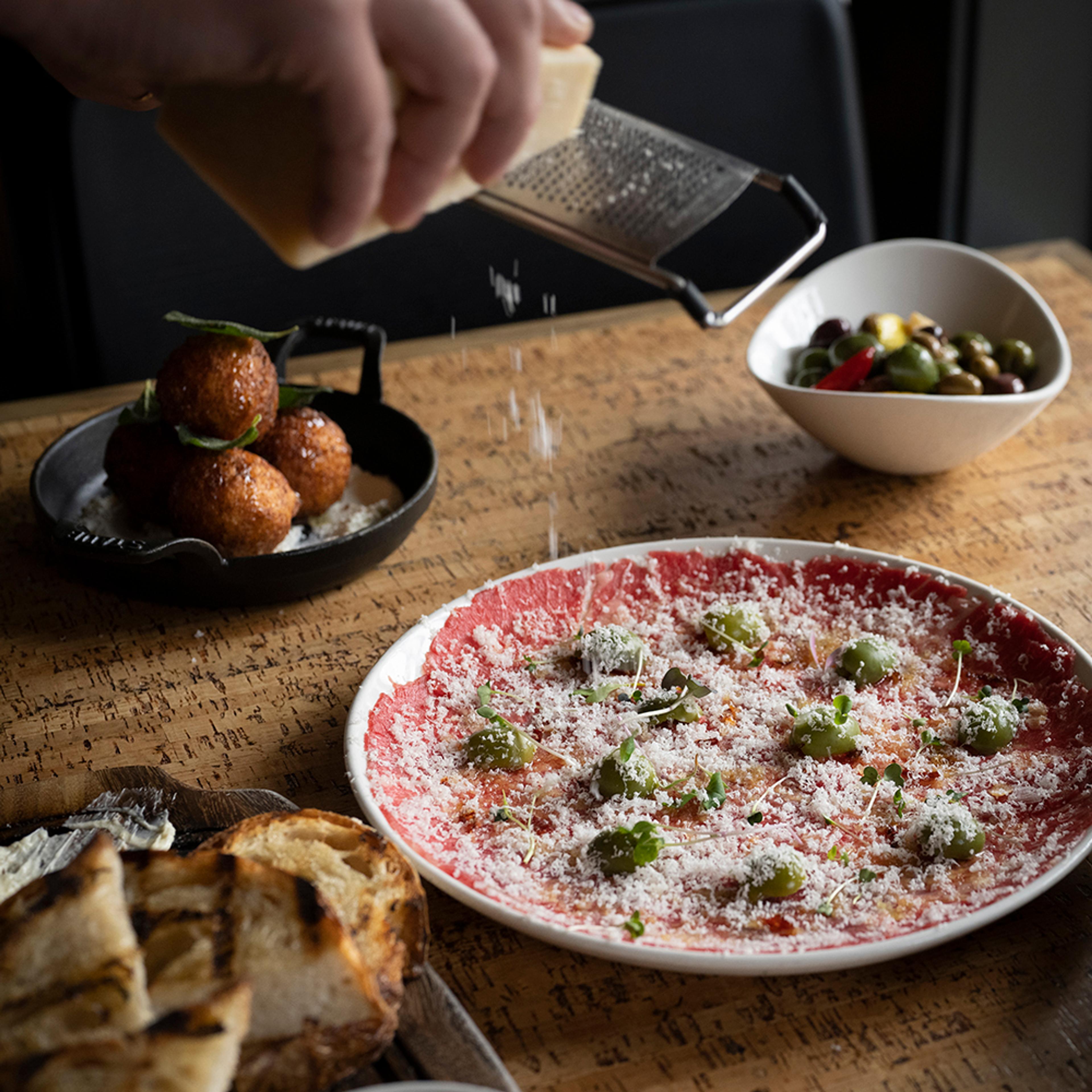 chef grating parmesan over beef carpaccio with pea vines