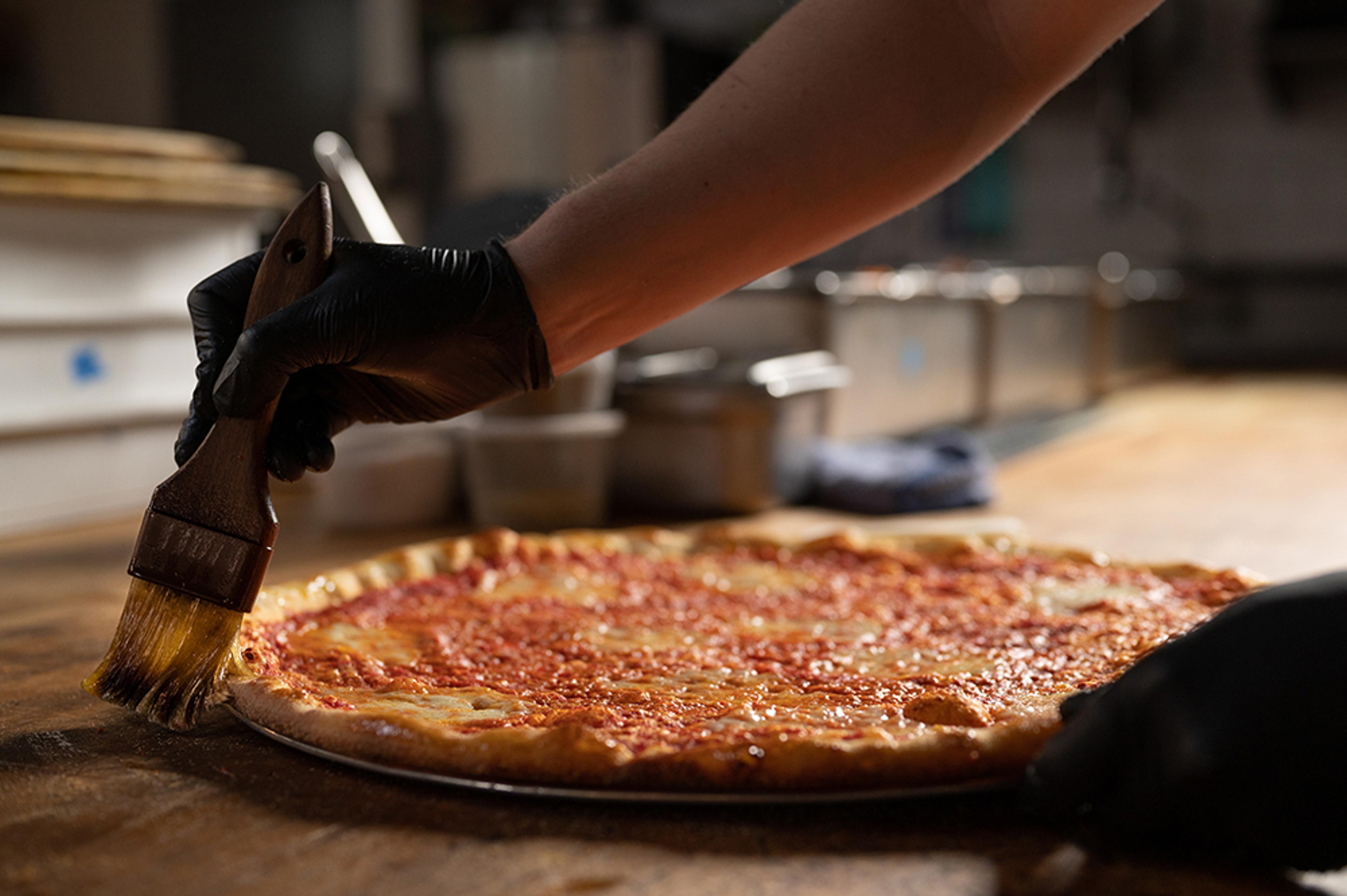 chef's hand applying olive oil to crust on a cheese pizza