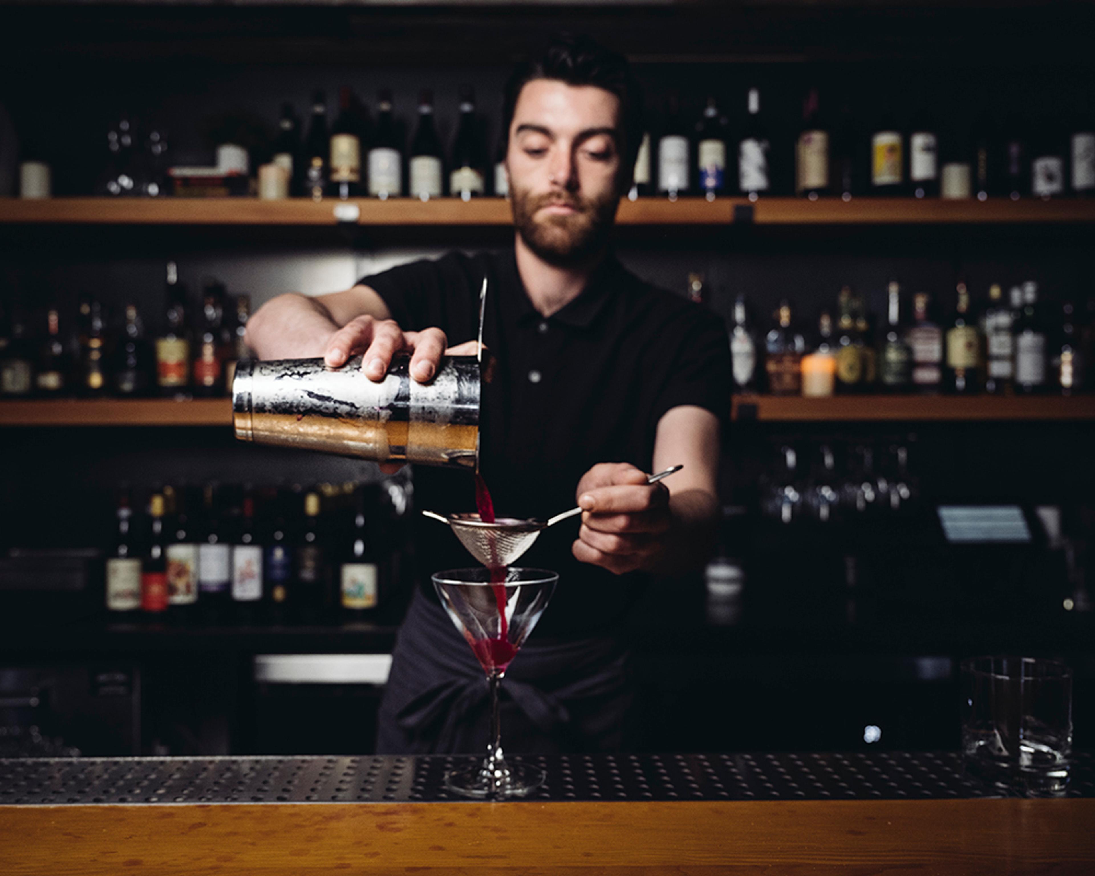 Bartender straining a deep purple cocktail into a martini stem glass