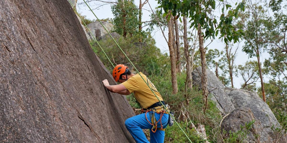 A climber in an orange helmet starting up a granite slab on a top rope, with gum trees and boulders behind.