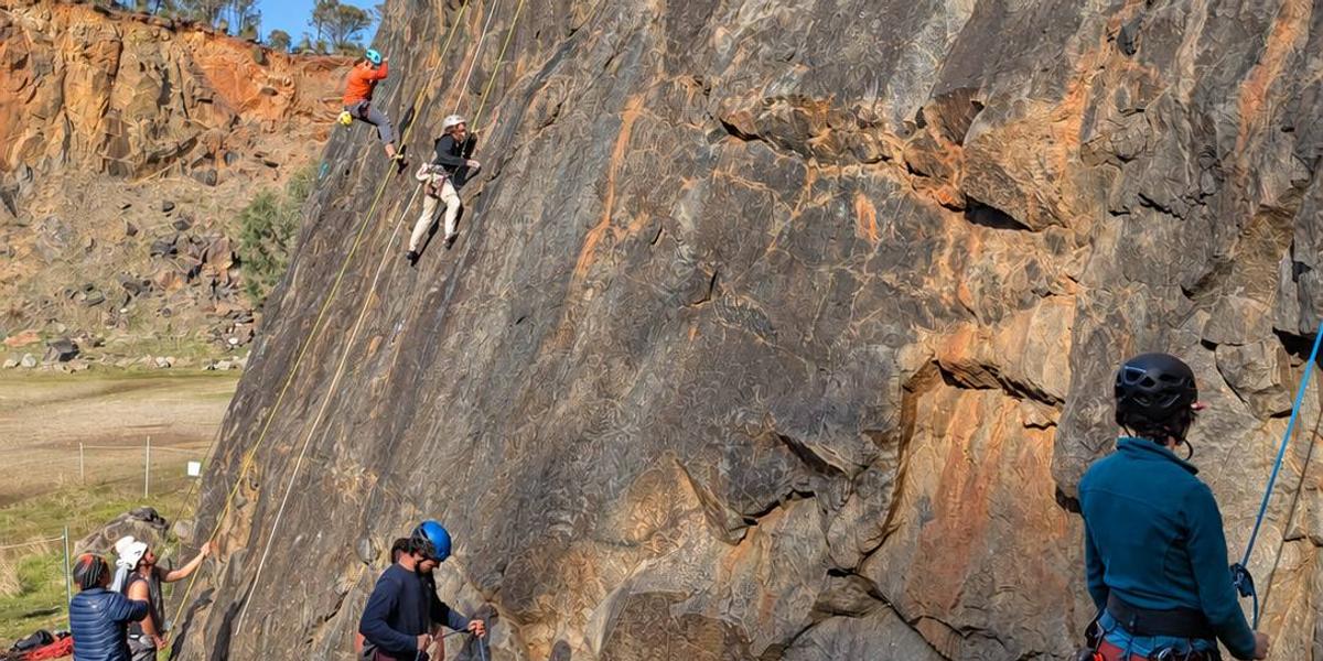 A group session at a sunny quarry wall: two climbers on top ropes and belayers at the base.